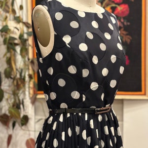 May include: A black and white polka dot dress with a black belt. The dress has a round neckline and is sleeveless. The polka dots are large and have a slightly raised texture.