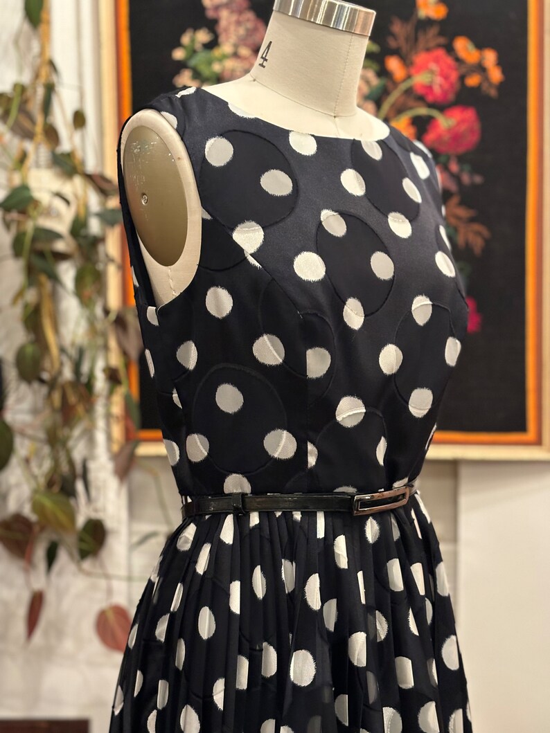 May include: A black and white polka dot dress with a black belt. The dress has a round neckline and is sleeveless. The polka dots are large and have a slightly raised texture.