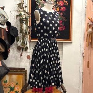 May include: A black and white polka dot dress with a pleated skirt. The dress is sleeveless and has a round neckline. It is on a mannequin in a shop.