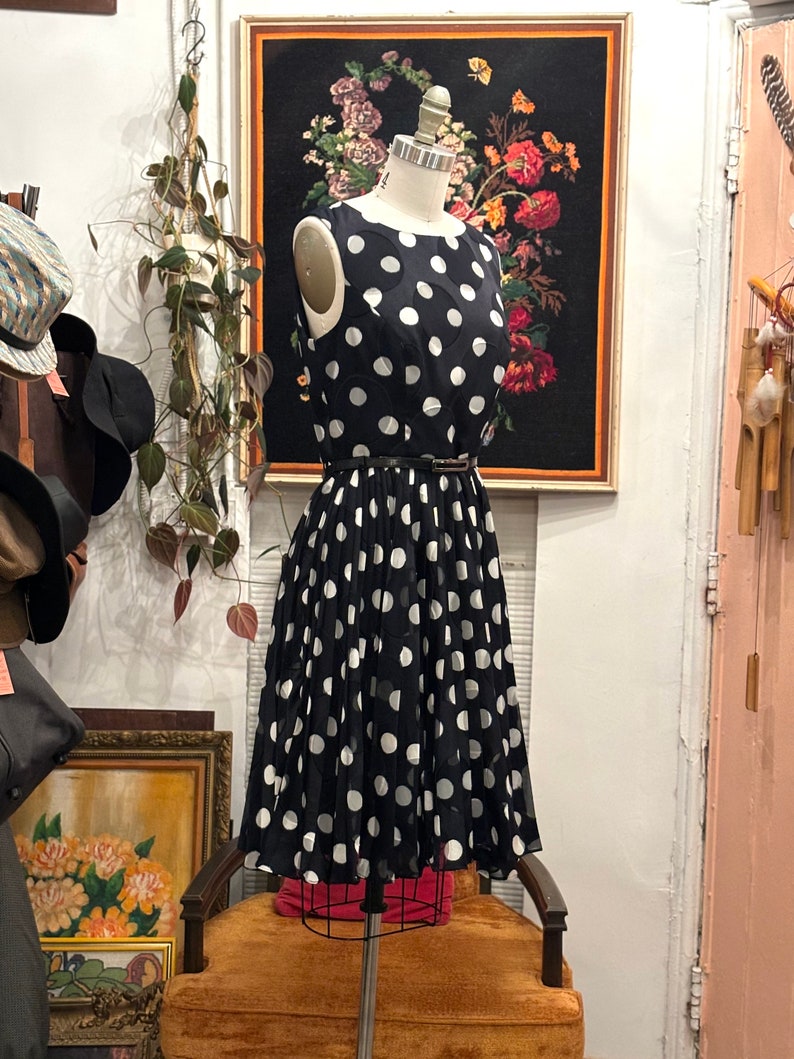 May include: A black and white polka dot dress with a pleated skirt. The dress is sleeveless and has a round neckline. It is on a mannequin in a shop.