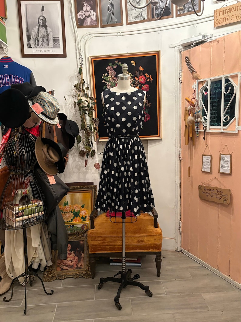 May include: A black and white polka dot dress on a mannequin in a shop. The dress has a fitted bodice and a full skirt. The mannequin is standing on a rolling stand in front of a yellow chair.