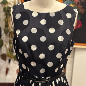 May include: A sleeveless black and white polka dot dress with a black belt. The dress has a round neckline and a pleated skirt.