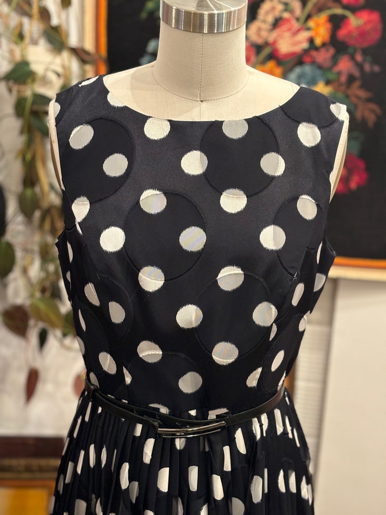 May include: A sleeveless black and white polka dot dress with a black belt. The dress has a round neckline and a pleated skirt.