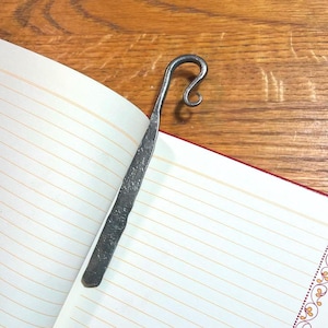 Puede incluir: Un marcapáginas artesanal de metal oscuro con una parte superior curvada y un acabado texturizado. El marcapáginas está insertado en un cuaderno rayado con detalles decorativos de borde naranja y rojo.