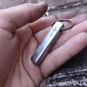 May include: A silver-colored rectangular keychain pendant with a cross and vertical line design. The pendant is attached to a brass-colored ring. The item is held in a hand.