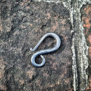 Puede incluir: Un gancho de metal plateado con un diseño único en forma de S. El gancho está hecho a mano y tiene un aspecto rústico e industrial.