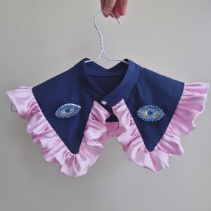 May include: A navy blue and pink ruffled collar with two blue eye-shaped brooches. The collar is held up by a white hanger. The collar has a button closure.