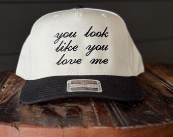 May include: White baseball cap with a black brim. The front of the cap has black embroidered text that reads "you look like you love me".