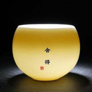 May include: A softly glowing, yellow-toned glass bowl with a rounded shape. The bowl has Chinese characters and a red seal. The interior is illuminated, creating a warm, inviting ambiance. The bowl is set against a dark background.