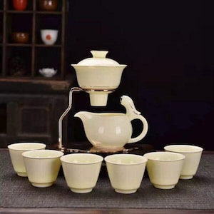 May include: A cream-coloured tea set comprising a teapot, a strainer, and six small cups. The teapot features a distinctive handle design. The set is arranged on a dark surface, suggesting a focus on tea ceremony or presentation.