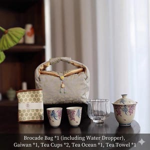 Può includere: Un set da tè con una borsa in broccato, gaiwan, tazze da tè, oceano da tè e un canovaccio. Le tazze da tè presentano un motivo floreale rosa e blu. La borsa è grigio chiaro con interno e manico dorati. Il testo sull'immagine recita: Brocade Bag *1 (including Water Dropper), Gaiwan *1, Tea Cups *2, Tea Ocean *1, Tea Towel *1