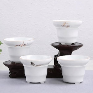 May include: Four white ceramic teacups with a flared rim and small feet. Each cup has a gold rim and a delicate painted design. The cups are displayed on dark wooden stands and a gray surface.