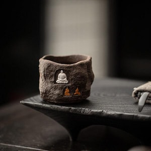 May include: A small, textured brown ceramic cup with a rough, stone-like appearance. The cup features three embossed Buddha figures in white and gold. The cup is sitting on a dark wooden surface.