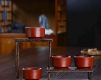 May include: Four small, reddish-brown ceramic tea cups are displayed on a tiered wooden stand. The cups have a simple, rounded design. The stand is made of dark wood and has a rustic appearance. The background includes a wooden cabinet and a dark surface.