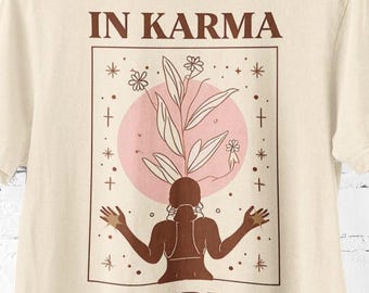 Puede incluir: Camiseta color crema con un gráfico marrón. El gráfico presenta una figura con los brazos extendidos, un círculo rosa e ilustraciones de plantas. El texto "IN KARMA WE TRUST" está impreso arriba y abajo del gráfico.