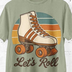 May include: Sage green t-shirt with a retro roller skate design. The graphic features a cream-coloured skate with brown accents and orange wheels, set against a sunset-style background. The text "Let's Roll" is below the skate.