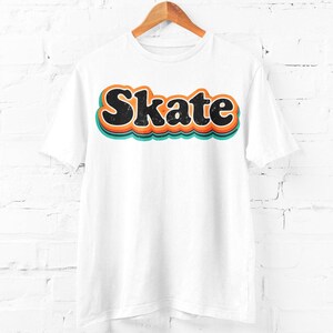 Peut inclure: T-shirt blanc avec le mot "Skate" en police r&eacute;tro. Le mot est noir avec des contours orange et turquoise, cr&eacute;ant un effet de superposition. Le t-shirt est suspendu &agrave; un cintre en bois devant un mur de briques blanches.