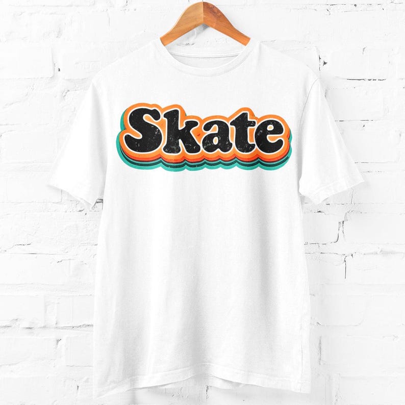 Peut inclure: T-shirt blanc avec le mot "Skate" en police r&eacute;tro. Le mot est noir avec des contours orange et turquoise, cr&eacute;ant un effet de superposition. Le t-shirt est suspendu &agrave; un cintre en bois devant un mur de briques blanches.