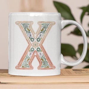 May include: White ceramic mug with a floral design in shades of green, pink, and brown. The letter "X" is featured in the center of the mug.