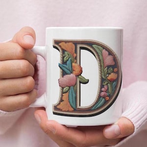May include: White ceramic mug with a floral embroidered letter D. The letter is outlined in black and filled with a variety of colors, including pink, orange, and green.
