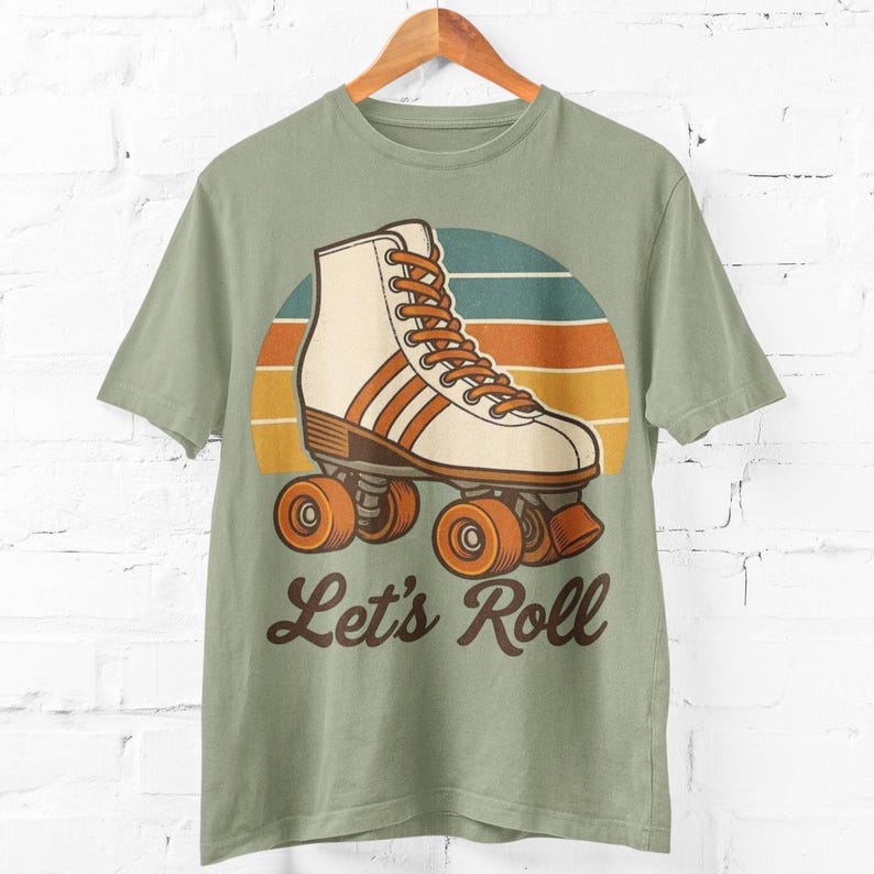 May include: Sage green t-shirt with a retro roller skate design. The graphic features a cream-coloured skate with brown accents and orange wheels, set against a sunset-style background. The text "Let's Roll" is below the skate.