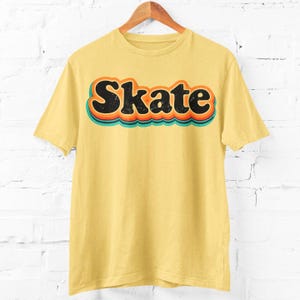 Peut inclure: T-shirt jaune clair avec le mot "Skate" dans une police r&eacute;tro. Le mot est noir avec des contours orange, turquoise et rouge. Le t-shirt est suspendu &agrave; un cintre en bois sur un mur de briques blanches.
