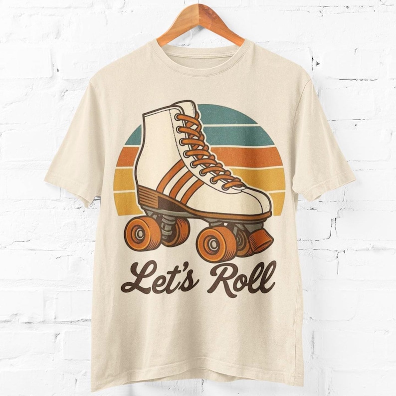 May include: A cream-coloured t-shirt with a retro roller skate design. The graphic shows a white and orange roller skate with orange wheels, set against a striped background of orange, yellow, and teal. The text "Let's Roll" is below the skate.