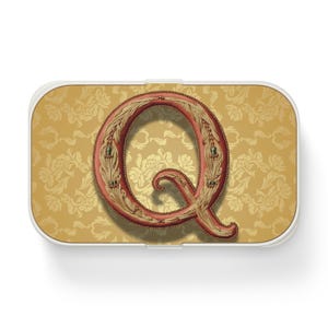 Könnte beinhalten: Eine weiße rechteckige Box mit einem gold- und braunen gemusterten Hintergrund. Der Buchstabe Q befindet sich in der Mitte der Box, mit einem roten und goldenen Design.