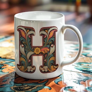 Puede incluir: Taza de cerámica blanca con un diseño floral colorido con la letra "H" en el centro.