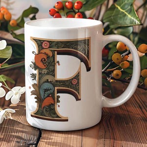 Può includere: Tazza in ceramica bianca con una lettera "F" decorativa e ornata in tonalità di verde, blu, arancio e oro. La lettera è circondata da motivi floreali e foglie.