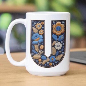 May include: White ceramic mug with a floral design in shades of blue, gold, and white. The letter "U" is formed by the floral design.