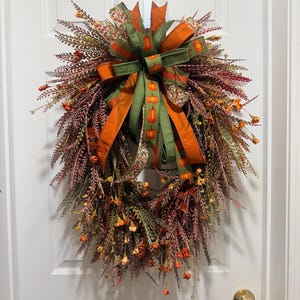 May include: A teardrop-shaped autumn wreath featuring a mix of dark red, brown, and green foliage with small orange flowers. A large decorative bow sits at the top, made of solid orange ribbon and green ribbon with a repeating orange pumpkin print.