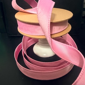 May include: Pink velvet ribbon spooled on a cardboard roll, with a length of ribbon cascading down. The ribbon is a soft, light pink color. The spool is sitting on a white pedestal. The background is dark.