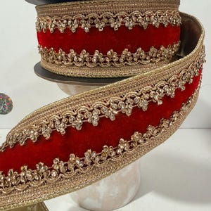 May include: A spool of wide ribbon featuring a deep red velvet center stripe bordered by intricate gold metallic trim. The edges are embellished with a repeating pattern of small, sparkling gold beads, creating a textured and ornate appearance.