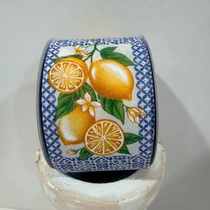 May include: A roll of wired ribbon featuring a white background with a blue geometric lattice border. The center pattern displays bright yellow lemons, green leaves, and small white blossoms, creating a vibrant and detailed botanical print.
