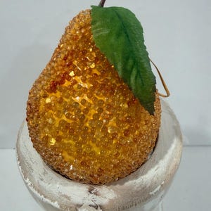 May include: A pear-shaped ornament covered in small, translucent amber-colored beads. The ornament features a single green fabric leaf attached to a brown stem at the top and includes a thin gold hanging string.