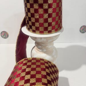 May include: A spool of decorative ribbon with a checkered pattern in burgundy and gold glitter. The ribbon is displayed on a white pedestal, with a length of the ribbon unspooled and visible.