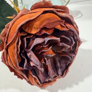 May include: A single artificial flower with layered fabric petals in shades of burnt orange and deep purple. The bloom features a dense, rounded shape with ruffled edges and a green stem with a single leaf extending from the side of the flower head.