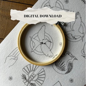 May include: A wooden embroidery hoop holds a white fabric with a black bird design. The fabric is part of a larger piece with other black line art designs, including a hand gesture, a bow and arrow, and the text "DIGITAL DOWNLOAD".