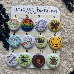 May include: A selection of twelve unique buttons for sale, each with a different design. The buttons feature various illustrations, including a dog, a smiley face, a butterfly, and text such as "PAWTASTIC" and "Certified Artist."