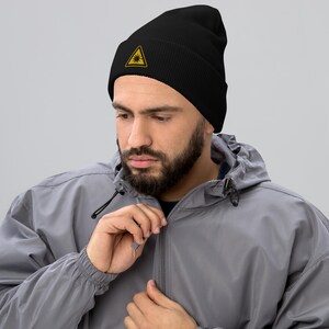 Op de afbeelding: Een zwarte beanie met een geborduurd geel driehoekig laserwaarschuwingssymbool aan de voorkant.