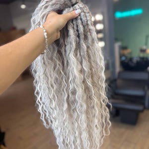 Puede incluir: Un conjunto de extensiones de cabello rizado de color gris claro. El cabello se sostiene en una mano, revelando la textura y las variaciones de color. Las extensiones tienen un aspecto natural y ondulado, con una mezcla de tonos grises claros y oscuros.