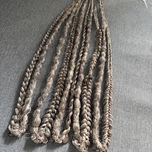 May include: Braided hair extensions in shades of brown and gray. The extensions are arranged in a fan shape, with multiple strands of braided hair. The braids are thick and textured, with a natural, earthy appearance. The hair extensions are designed for styling.