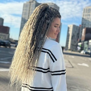 May include: A woman with long, blonde dreadlocks, styled in a high ponytail, is seen from the back. She wears a white long-sleeve shirt with black stripes across the chest and sleeves. The background features city buildings and a blue sky.