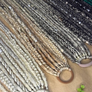 May include: Close-up of several braided hair extensions in various shades of gray, blonde, and brown. The extensions are secured with neutral-colored hair ties. The image showcases a variety of colors and textures, suitable for creating different hairstyles.