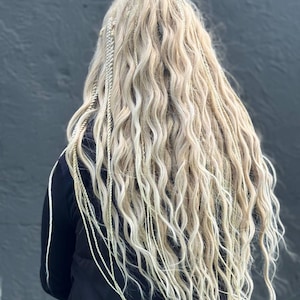 May include: A close-up of long, wavy, blonde hair with multiple thin, braided strands. The hair cascades down, creating a full, textured look. The style appears to be a form of hair extensions or a unique braiding technique.