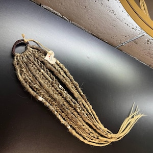 Può includere: Un set di dreadlock sintetici marroni e biondi, attaccati a un elastico marrone. I dreadlock presentano dettagli bianchi e una piccola etichetta a forma di cuore attaccata con spago. Su una superficie nera.