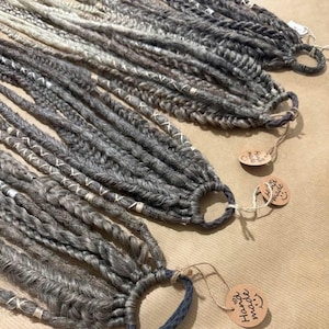 Op de afbeelding: Een verzameling grijze en witte dreadlocks, met de hand gebonden en versierd met witte draadaccenten. Elke dreadlockbundel heeft een lus aan het einde en een klein, rond label met de tekst "Hand made" met een smiley. De dreadlocks liggen op een lichtbruin oppervlak.