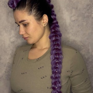 Op de afbeelding: Een vrouw met een lange, gevlochten haarstijl in paarse tinten. De vlecht is een opvallend kenmerk, die naar beneden over de rug valt. Ze draagt een olijfgroen shirt met lange mouwen met het woord "YES" herhaaldelijk gedrukt. De achtergrond is grijs.