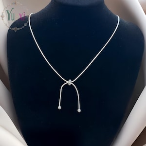 Puede incluir: Un delicado collar plateado con una cuenta deslizante y dos pequeños abalorios redondos en los extremos. El collar se exhibe sobre un maniquí negro, mostrando su diseño simple pero elegante. El logotipo de Yu-xi es visible en la esquina superior izquierda.